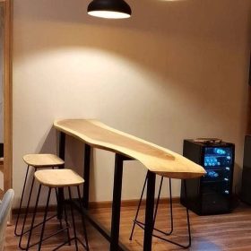 میز بار چوبی با لبه‌های طبیعی و پایه‌های فلزی مشکی، همراه با صندلی‌های بار چوبی و چراغ آویز مشکی در یک فضای مدرن.